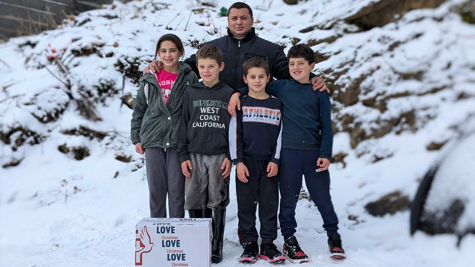 A family stand outside their home in the snow, smiling.