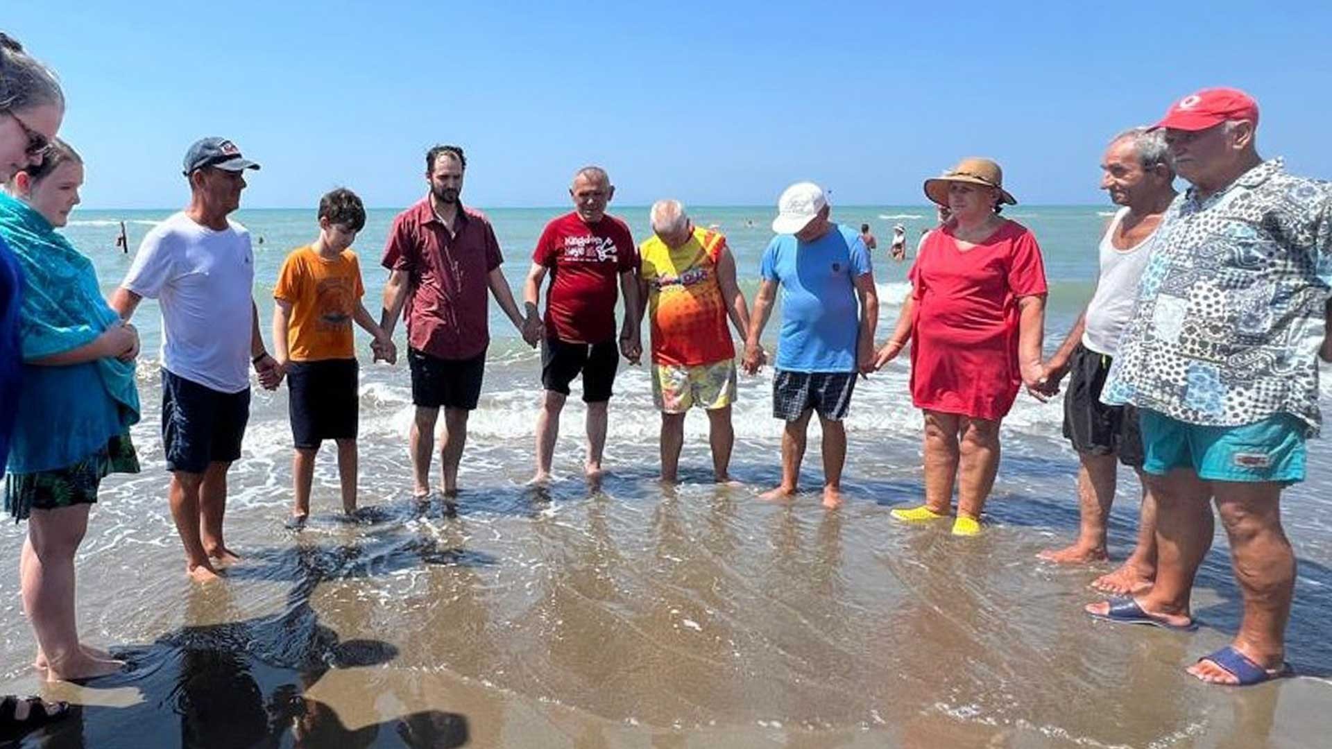 Group stands hand in hand in a half circle on the beach after a baptism.