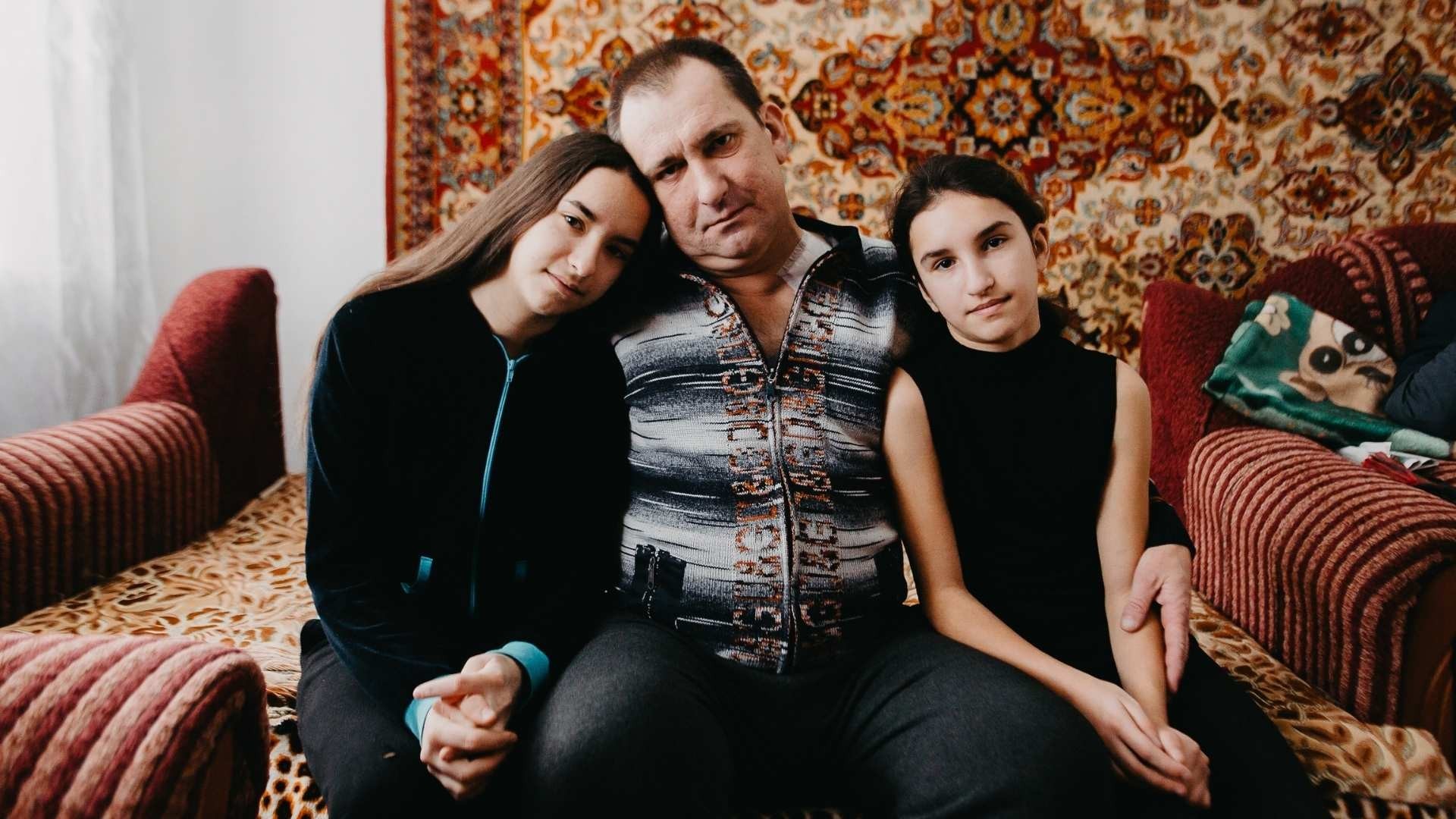 Father sits on a bed, holding his two daughters close