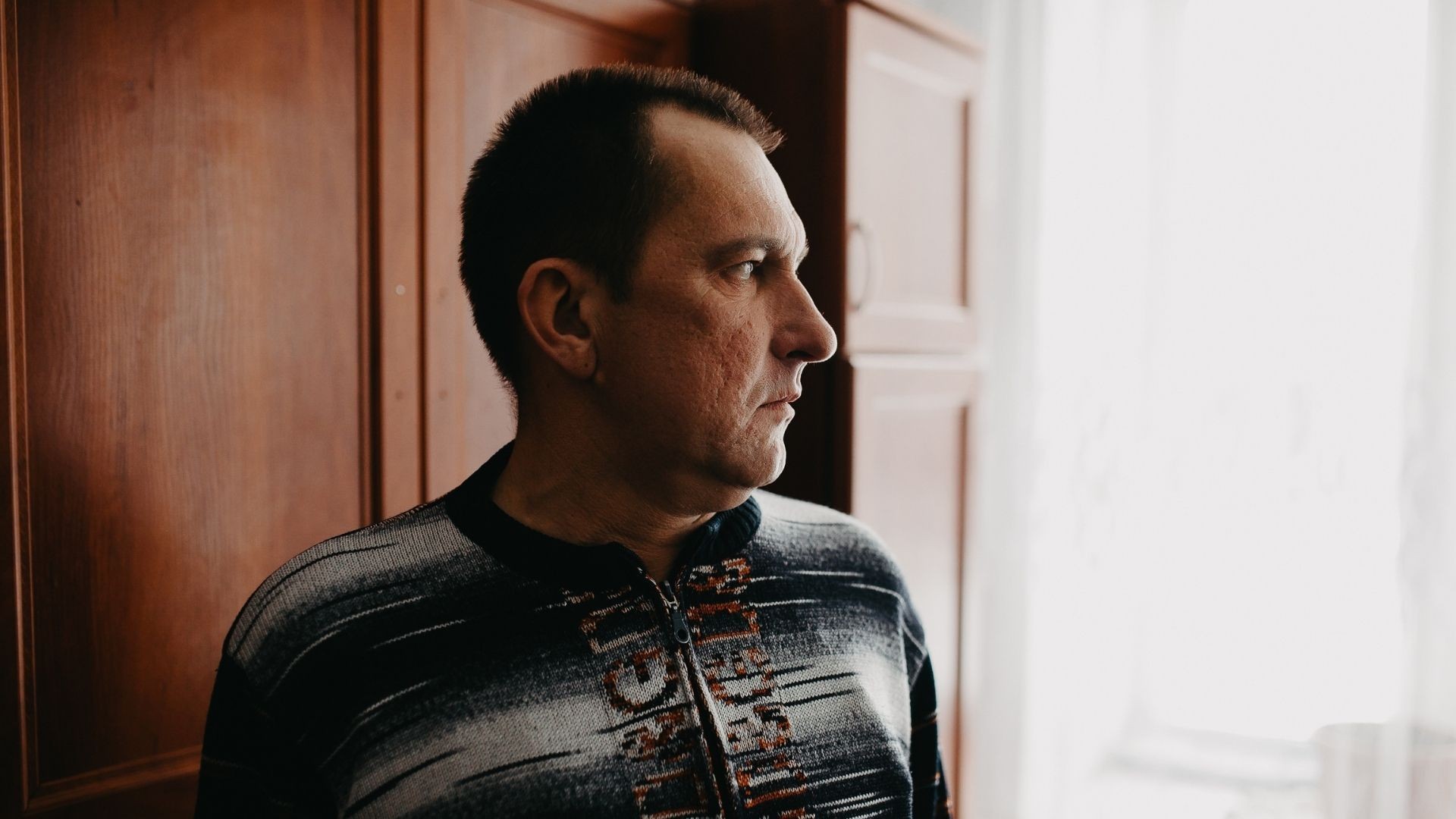 Man stands by a window, gazing out somberly, reflecting quiet grief and deep thought