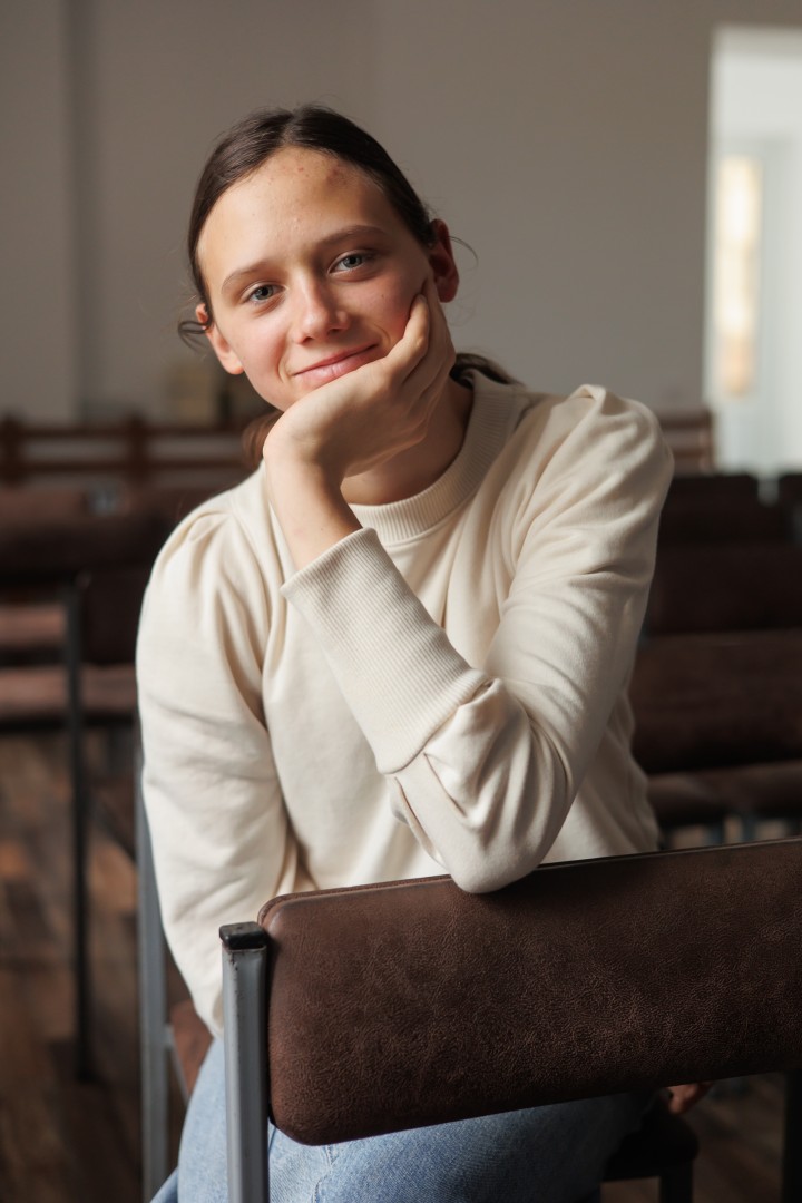 A girl leaning her elbow on a chair, her hand on her chin, smiling.